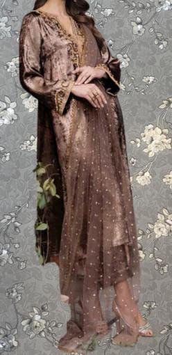 A woman wearing a long, brown dress with a sheer overlay and gold accents, standing against a gray floral background.