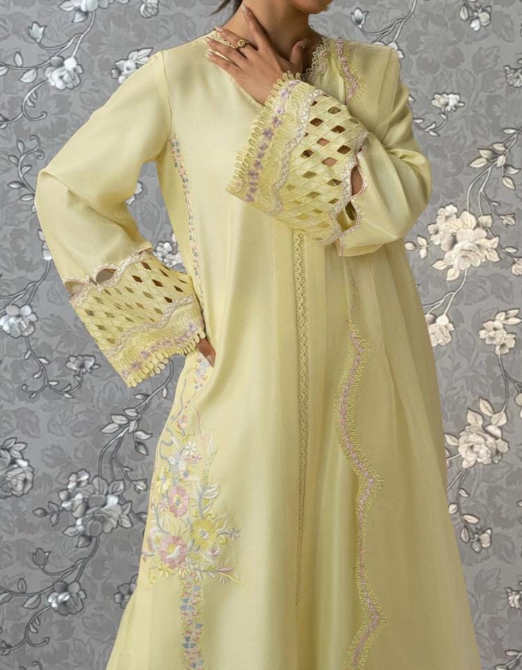 A woman wearing a long, yellow dress with intricate lace and floral embroidery, posing against a gray floral background.