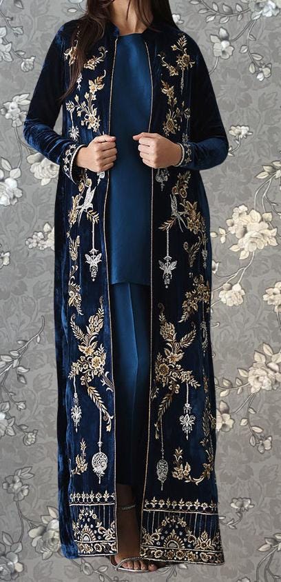 A woman wearing a long, blue velvet coat with gold embroidery stands against a gray floral background.