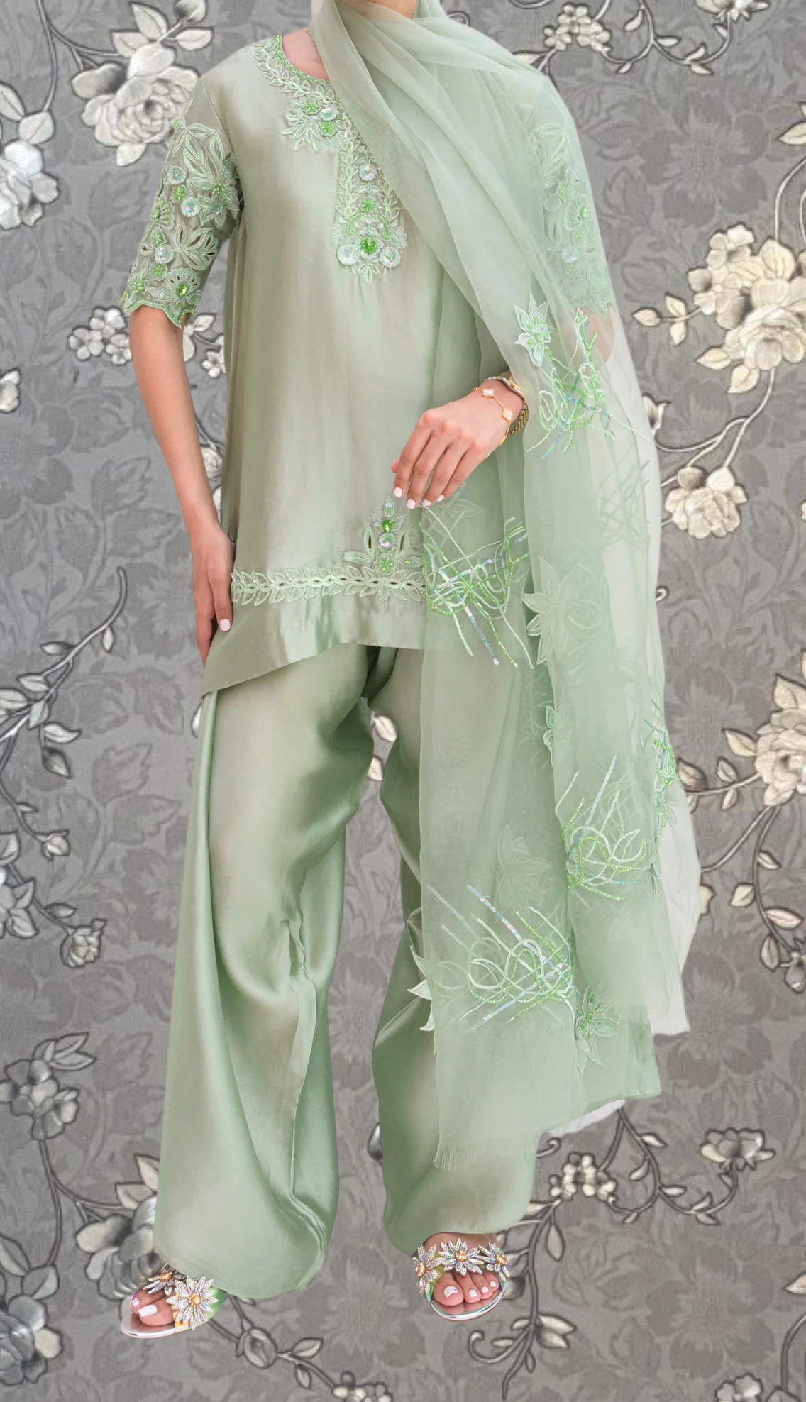 A person wearing a green dress with intricate embroidery and a matching green scarf, standing against a gray floral background.