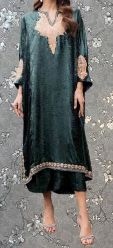 A woman wearing a long, green velvet dress with gold embroidery stands against a gray floral background.