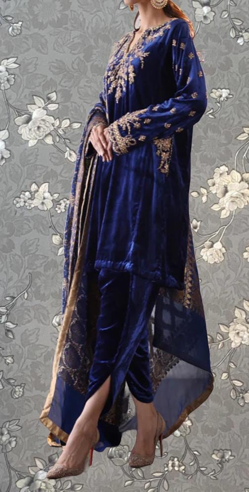 A woman wearing a blue velvet dress with gold embroidery and a matching shawl, standing against a gray floral background.