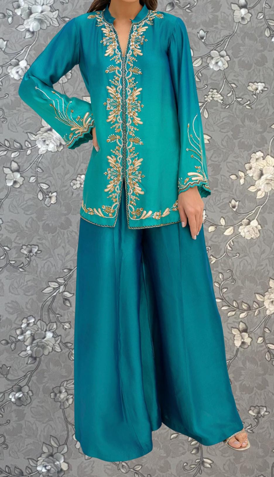 A woman wearing a turquoise and gold dress with intricate embroidery, standing against a gray floral background.