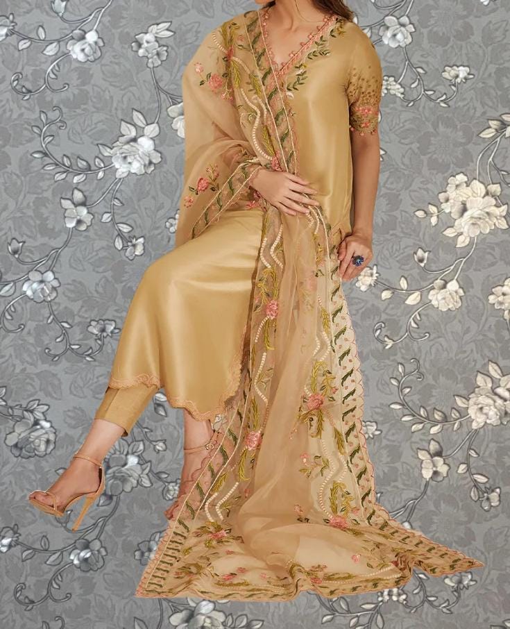 A woman wearing a gold and beige dress with intricate floral embroidery poses against a gray floral background.