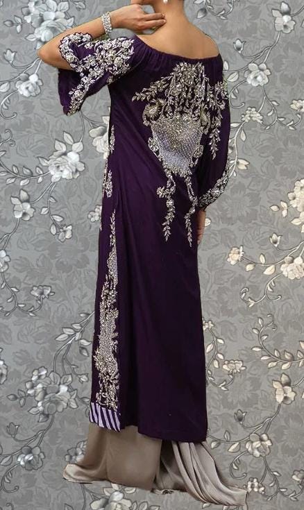 A woman wearing a long purple dress with intricate embroidery stands against a gray floral background.
