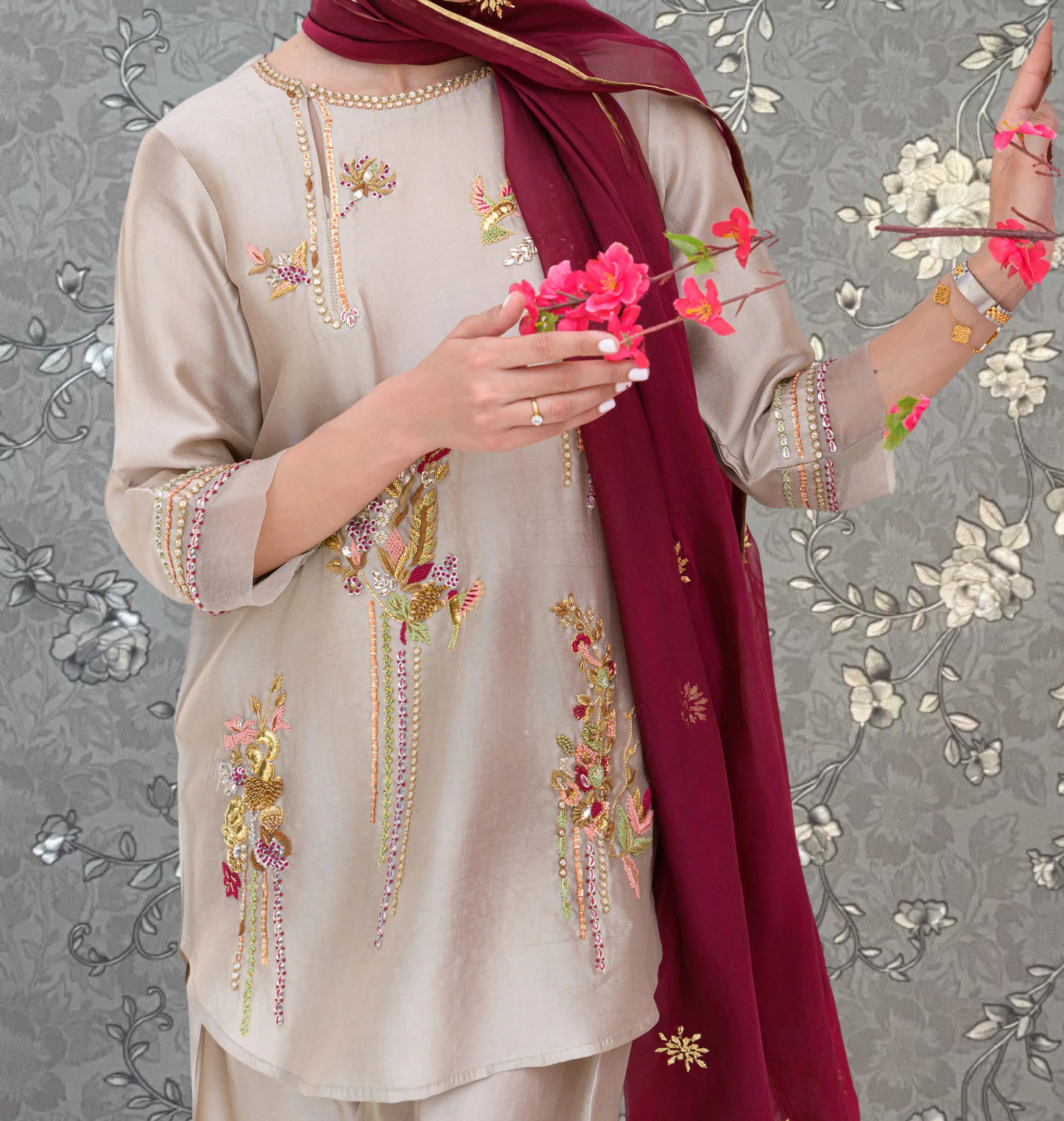 A person wearing a beige dress with intricate embroidery and holding a bouquet of pink flowers.