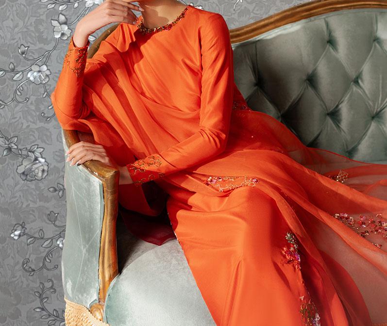 A woman wearing an orange dress with a long train sits on a gray and green upholstered chair against a gray floral wallpaper background.