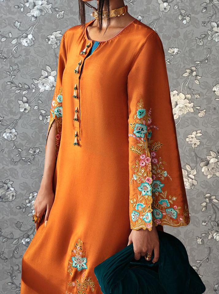 A woman wearing an orange dress with floral embroidery and a gold choker necklace stands against a gray floral background.