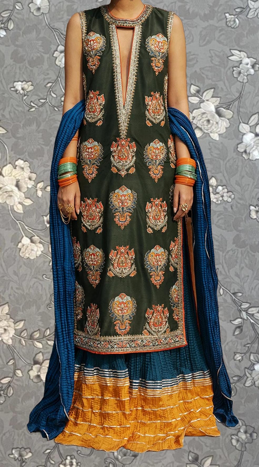 A woman wearing a long, sleeveless dress with a floral pattern and a matching blue shawl. She is also wearing multiple bracelets on her wrists.