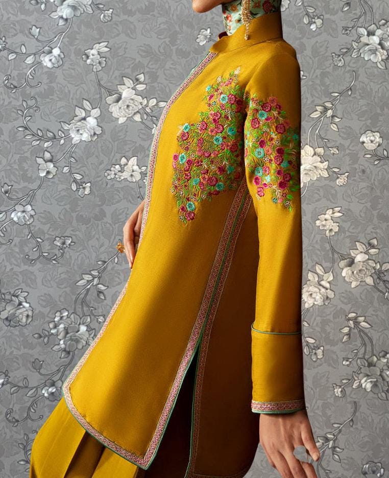 A person wearing a vibrant yellow dress with intricate floral embroidery on the sleeves and collar, posing against a gray floral background.
