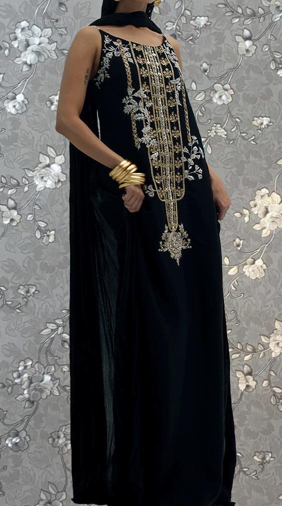 A woman wearing a long black dress with gold embroidery and jewelry poses against a gray floral background.
