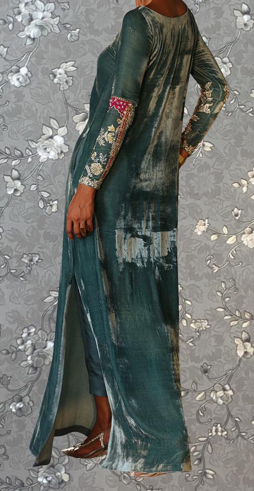 A person wearing a long, green dress with intricate embroidery and floral patterns, standing against a gray floral background.