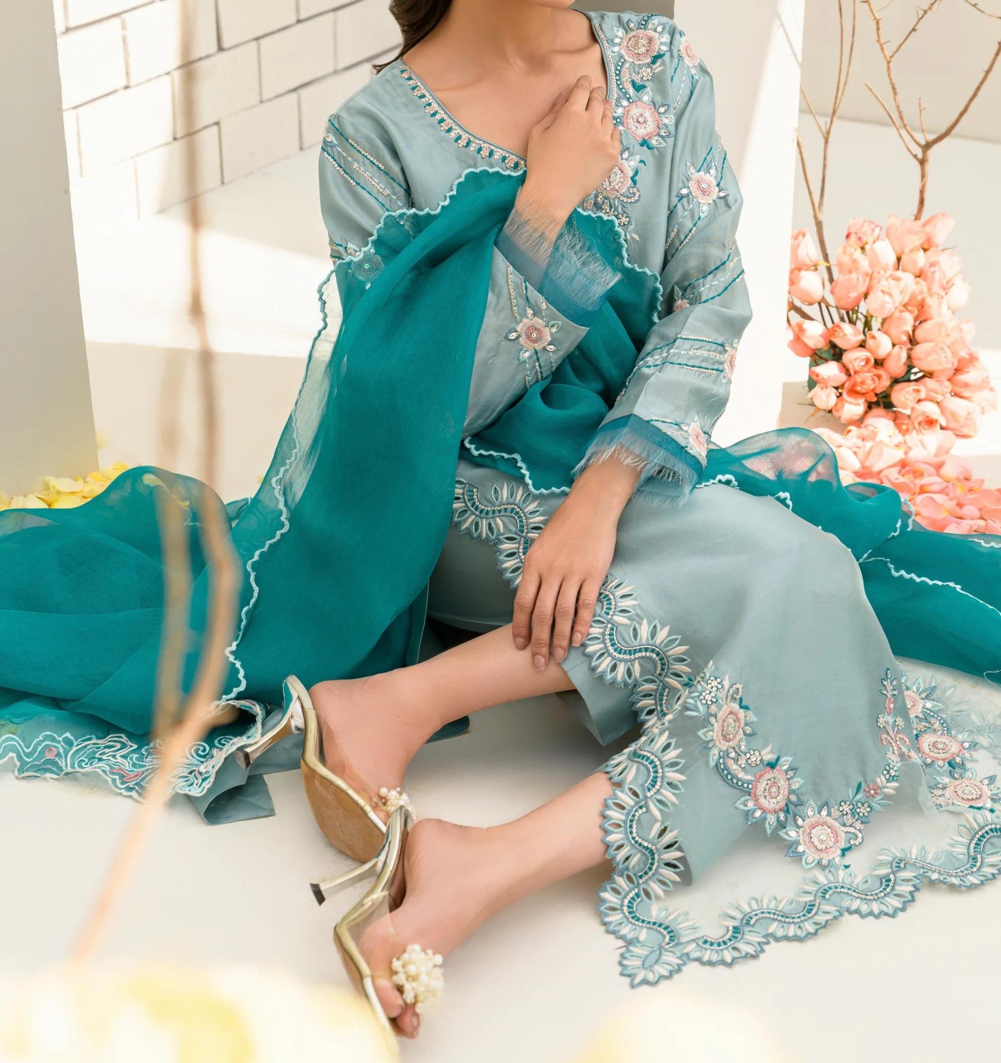 A woman wearing a teal and white dress with intricate embroidery sits on the floor, surrounded by pink flowers and a white wall.