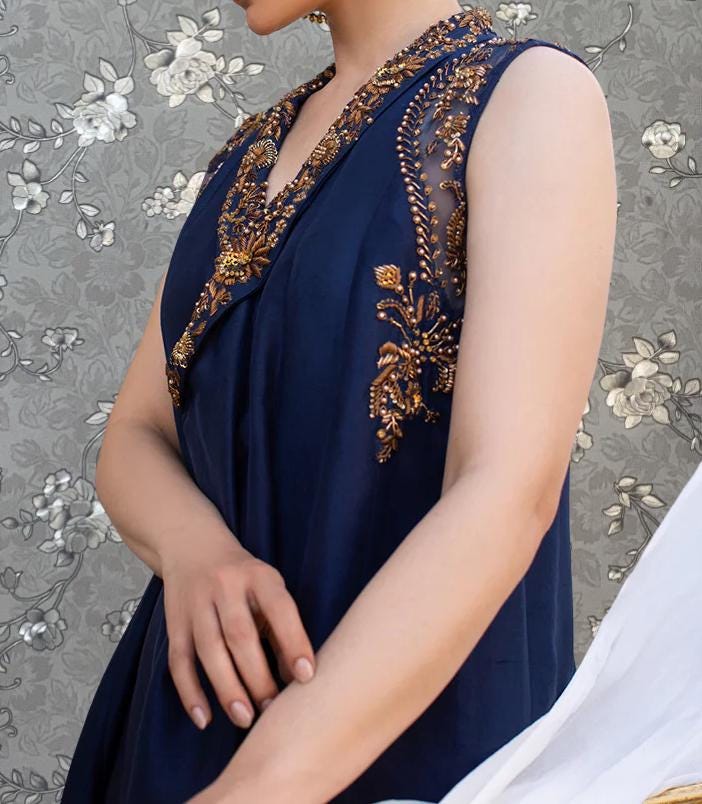 A woman wearing a navy blue dress with gold embroidery on the neckline and sleeves, posing against a gray floral background.