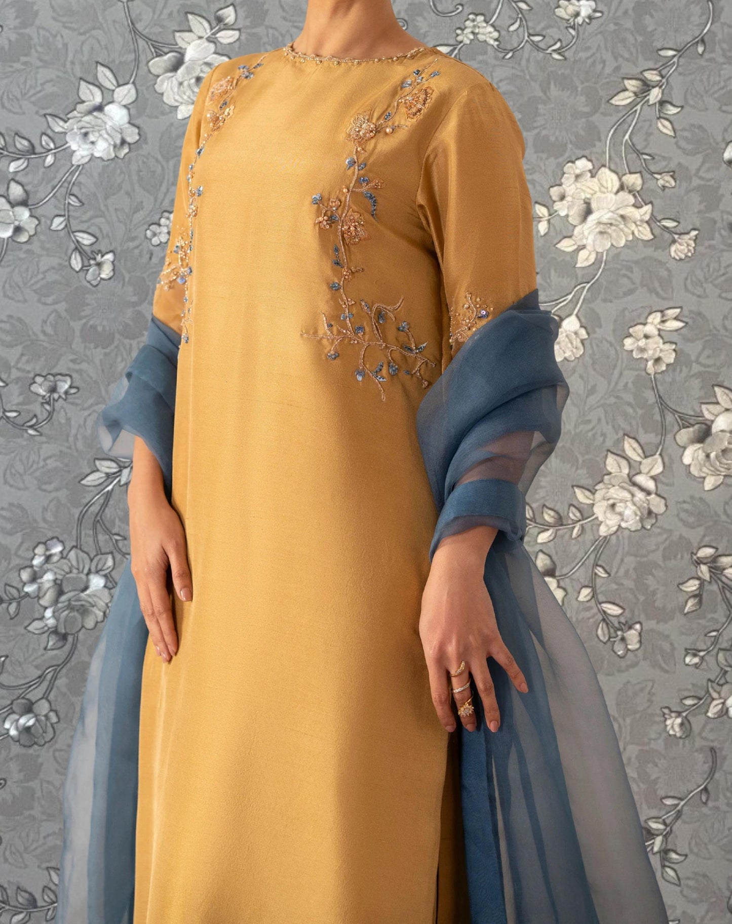 A person wearing a yellow dress with floral embroidery and a blue shawl stands against a gray floral background.