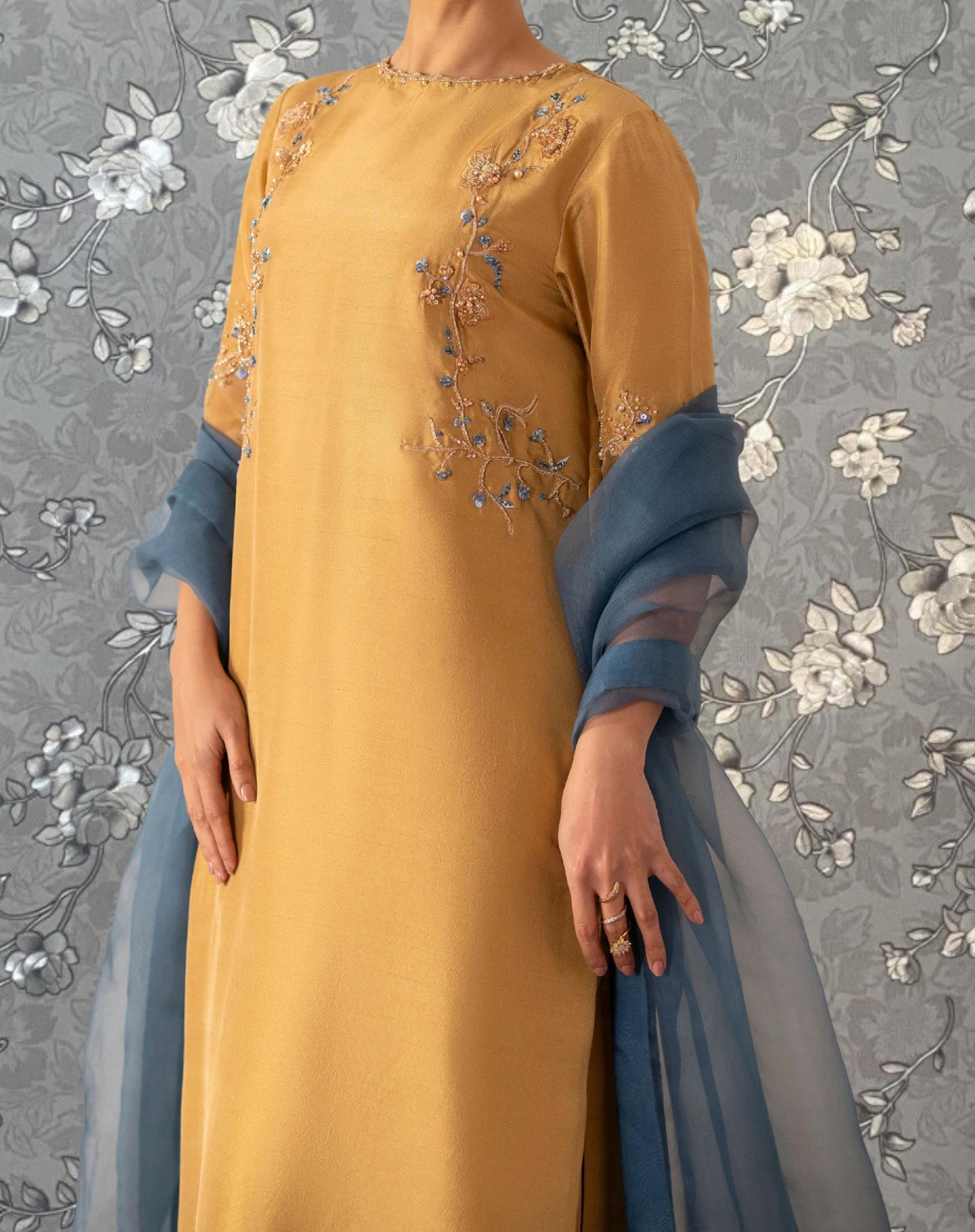 A person wearing a yellow dress with floral embroidery and a blue shawl stands against a gray floral background.