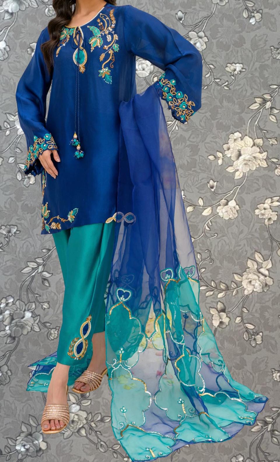 A woman wearing a blue dress with intricate embroidery and a matching blue and green skirt, standing against a gray floral background.