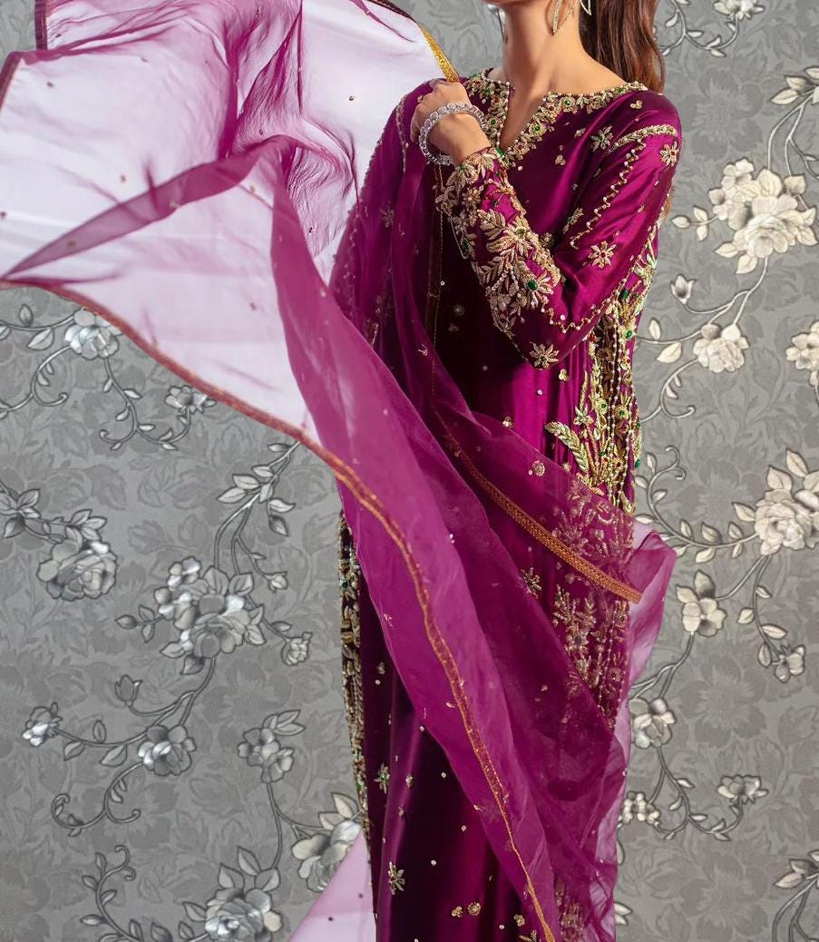A woman wearing a long, purple dress with gold embroidery and a matching purple shawl is standing against a gray floral background.
