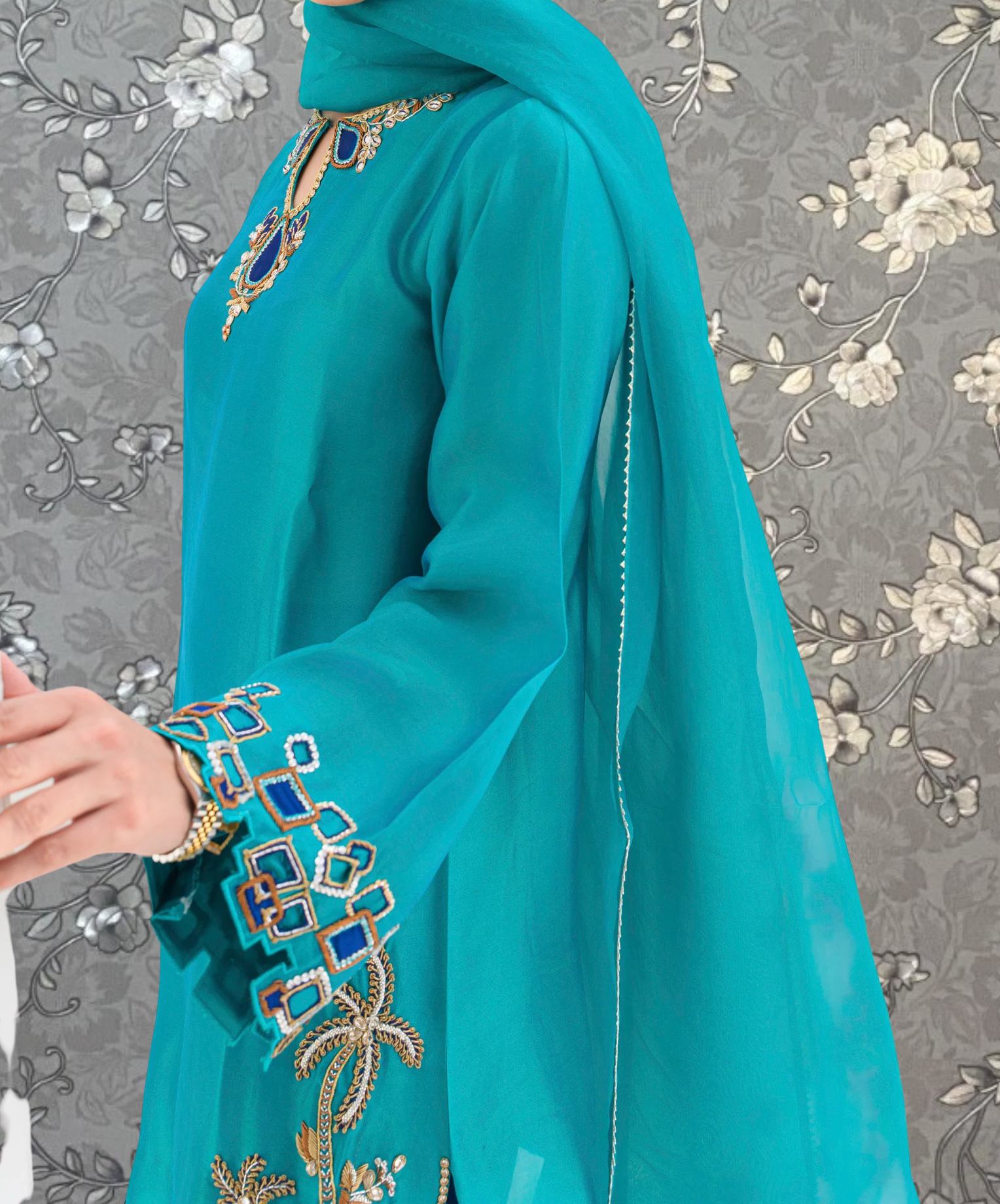 A person wearing a turquoise dress with intricate embroidered designs on the sleeves and collar, standing against a gray floral background.