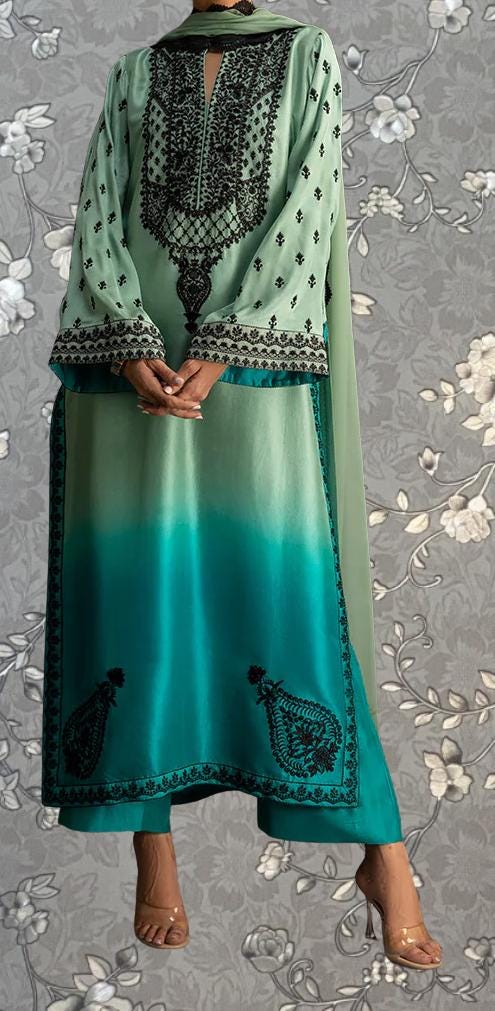 A person wearing a green and black patterned dress with intricate embroidery stands against a gray floral background.