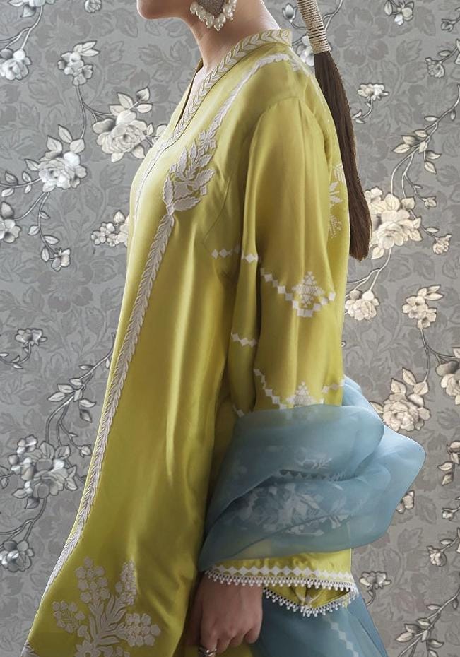 A woman wearing a yellow dress with white floral embroidery stands against a gray floral background.