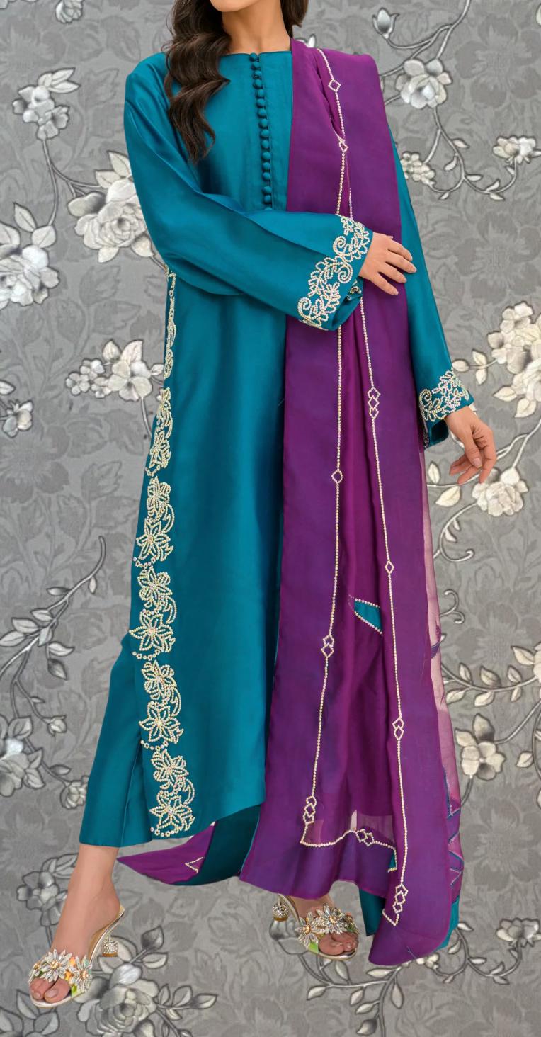 A woman wearing a long, teal-colored dress with a purple shawl draped over her shoulders, standing against a gray floral background.