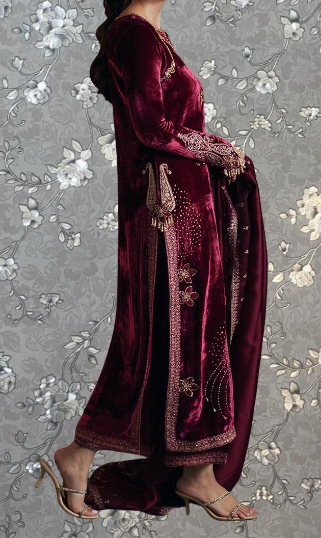 A woman wearing a long, burgundy velvet dress with intricate embroidery and a matching shawl. She is wearing high heels and posing against a gray floral background.