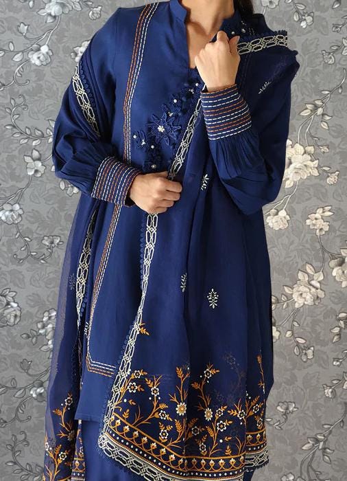 A woman wearing a blue dress with intricate embroidered patterns and designs, standing against a gray floral background.