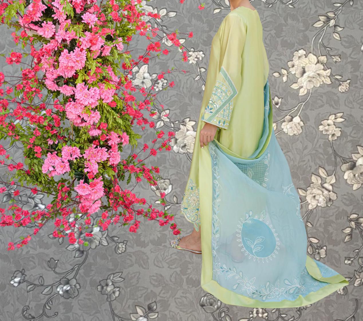 A person in a yellow dress stands in front of a floral arrangement with pink flowers, and a blue and white patterned fabric draped over their shoulder.