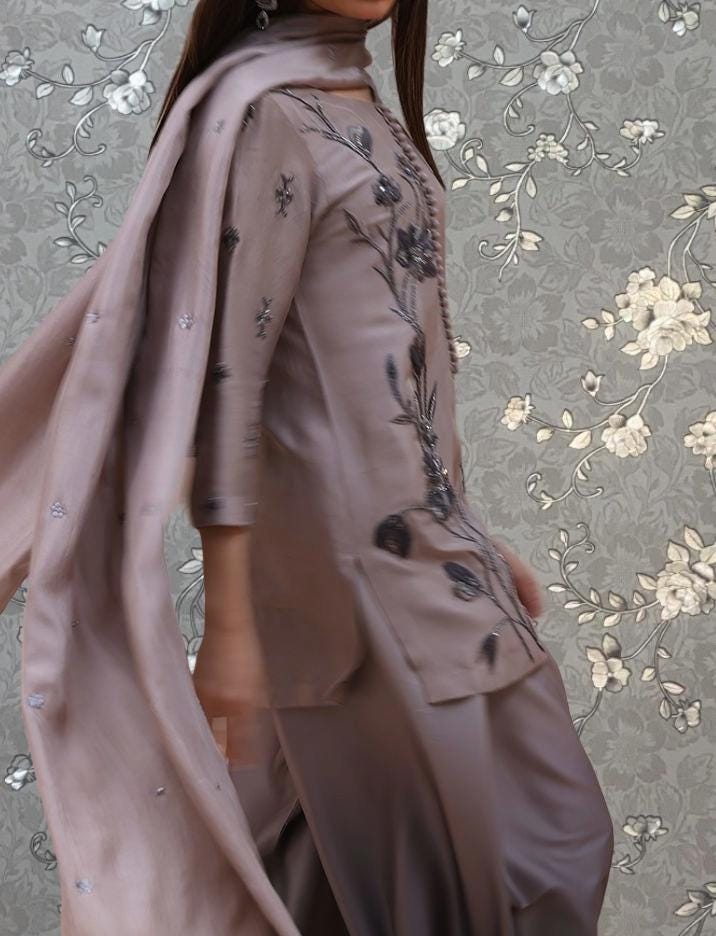 A woman wearing a brown dress with floral embroidery stands against a gray floral background.