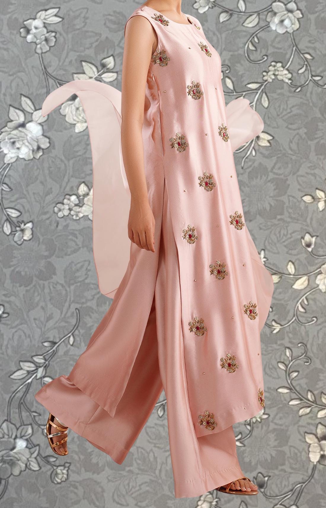 A woman wearing a pink dress with floral embroidery poses against a gray floral background.