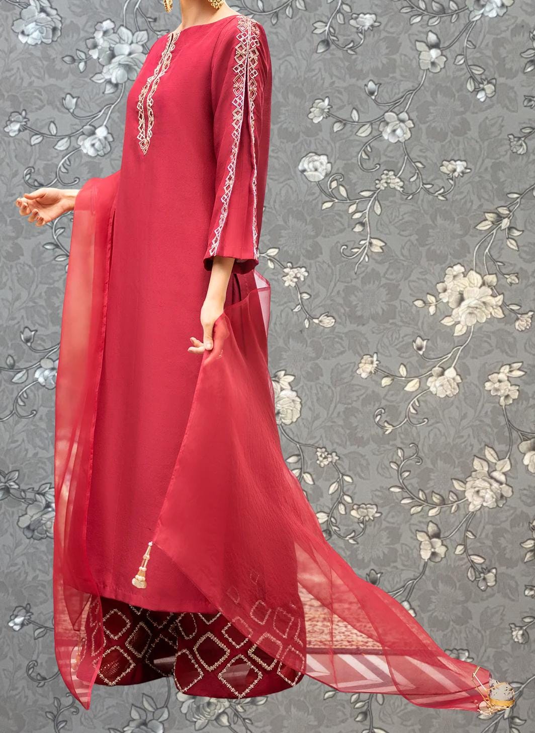 A woman wearing a long red dress with a matching red shawl, standing against a gray floral background.