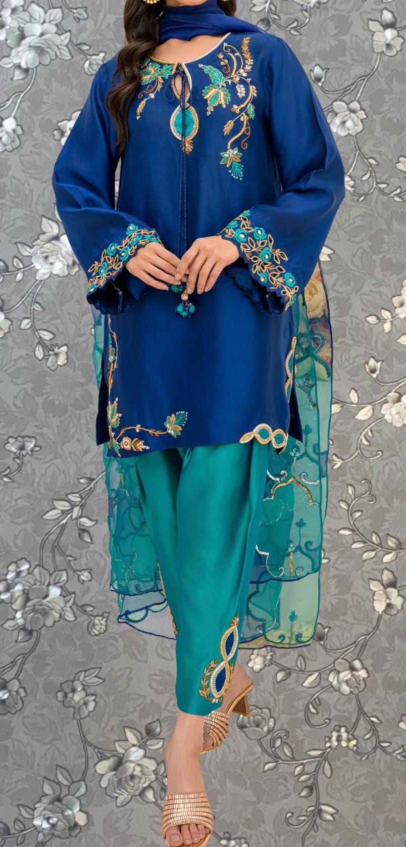 A woman wearing a blue dress with intricate embroidery and gold accents, paired with green pants, stands against a gray floral background.