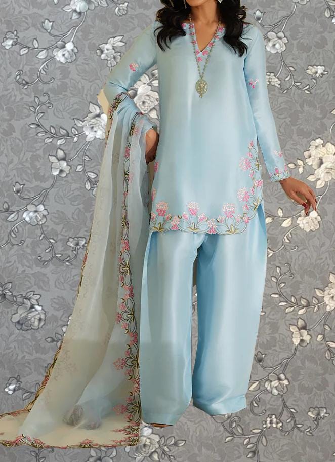 A woman wearing a light blue dress with pink floral embroidery, standing against a gray floral background.