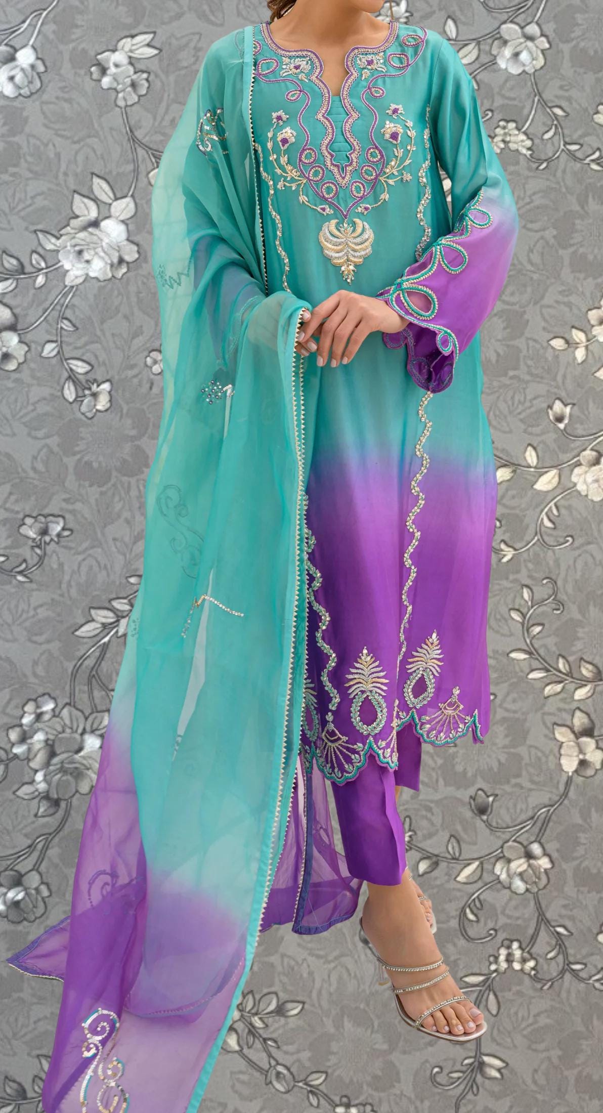 A woman wearing a colorful, patterned dress with a turquoise and purple color scheme, standing against a gray floral background.