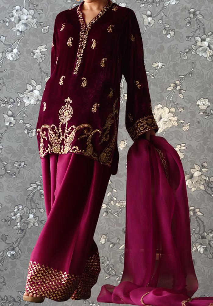 A person wearing a burgundy velvet dress with gold embroidery and a matching pink skirt, posing against a gray floral background.