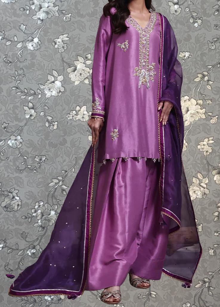 A woman wearing a purple dress with a matching purple shawl and a matching purple skirt, standing against a gray floral background.