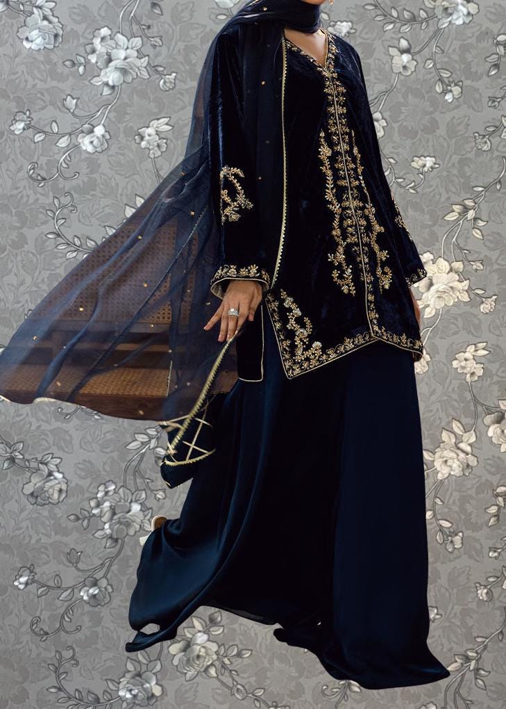 A woman wearing a long black dress with gold embroidery and a matching black veil, standing against a gray floral background.