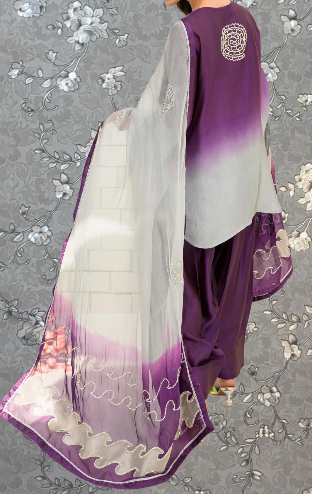 a person wearing a purple and white outfit with a long, flowing white shawl. The outfit includes a purple top and matching pants, and the person is standing against a gray floral background.