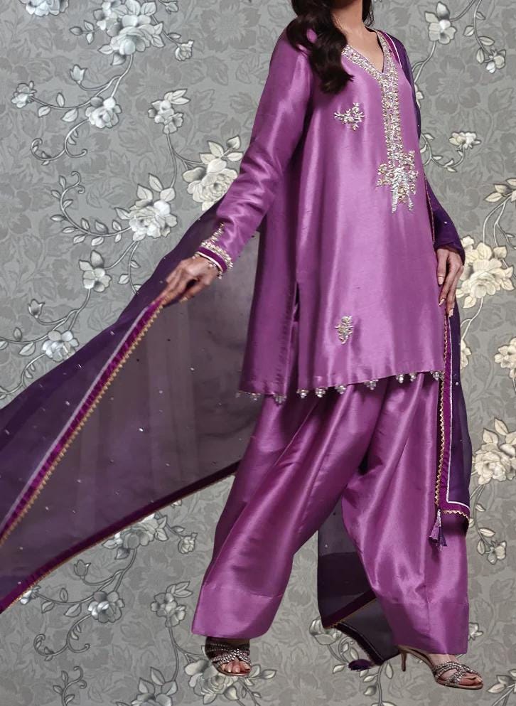 A woman wearing a purple dress with intricate embroidery and a matching purple shawl, standing against a gray floral background.