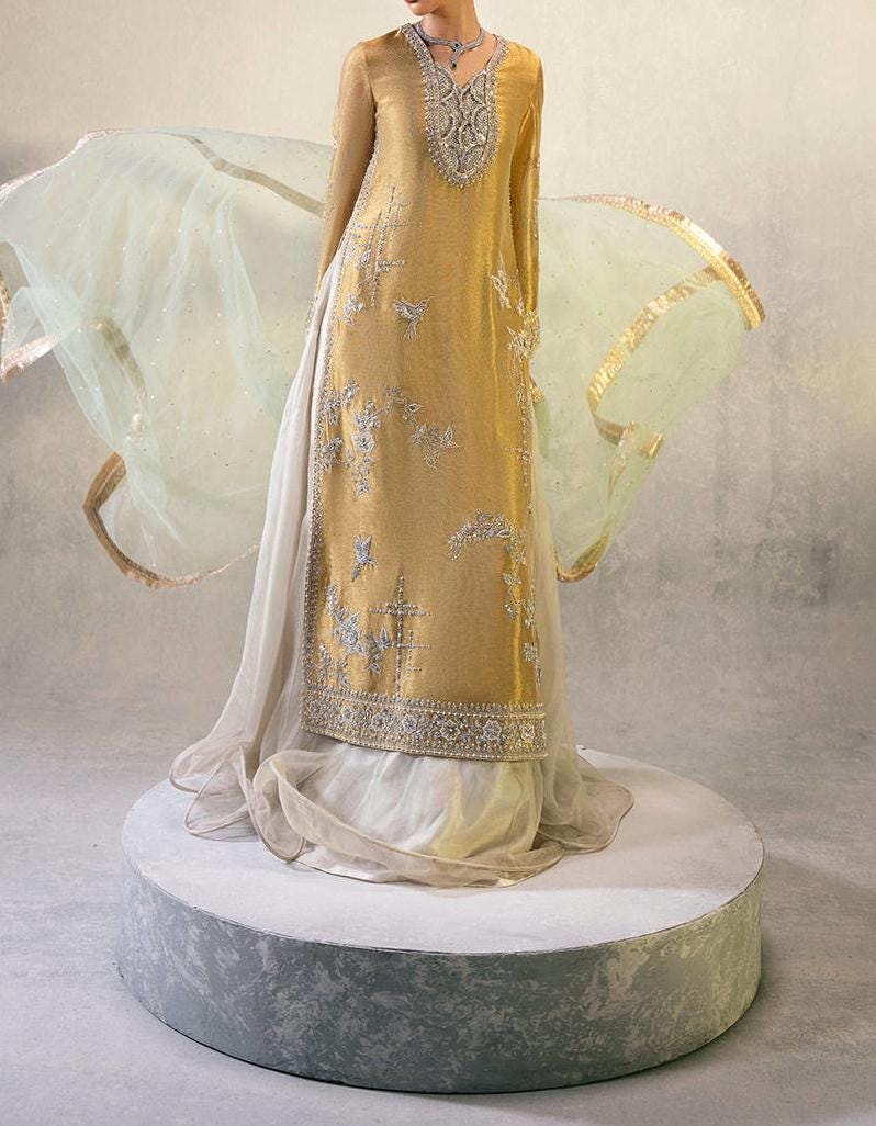 A woman wearing a gold and white dress with intricate embroidery stands on a pedestal, with her long white skirt flowing behind her.