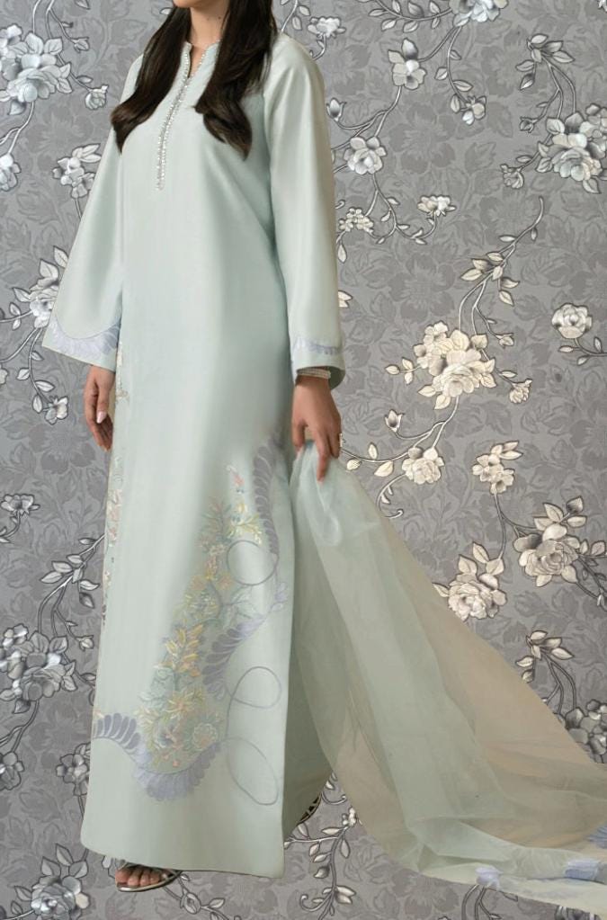 A woman wearing a long, light blue dress with floral embroidery stands against a gray floral background.