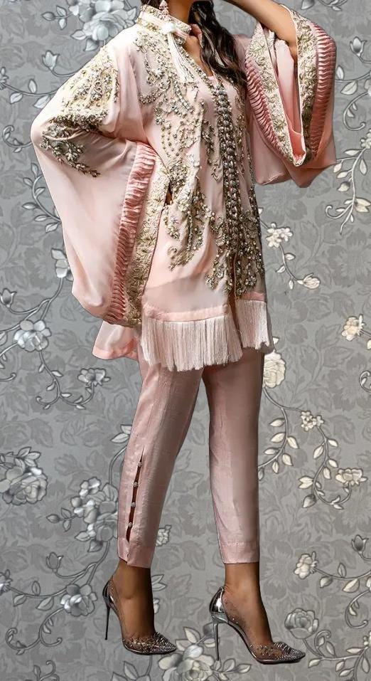 A woman wearing a pink and gold dress with intricate embroidery and frills, standing against a gray floral background.