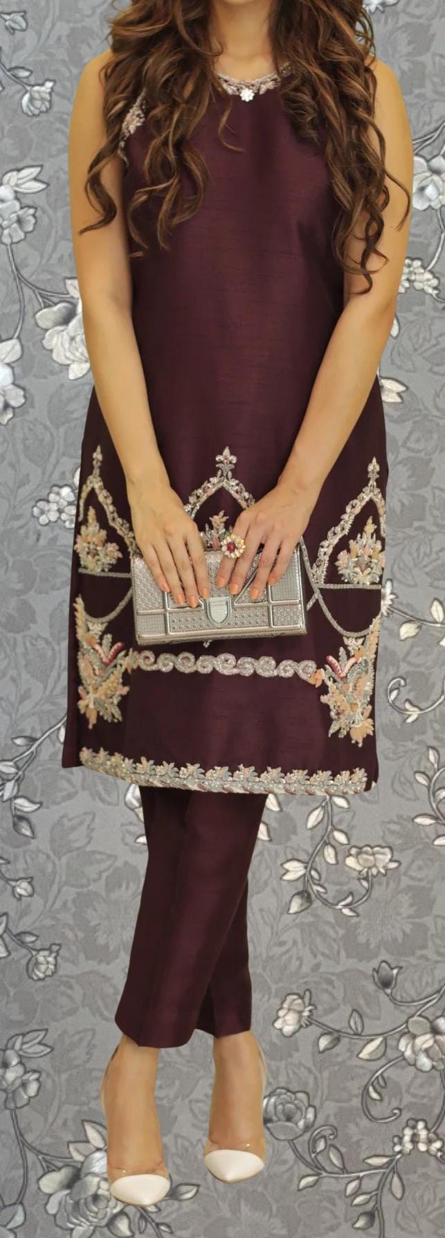 A woman wearing a burgundy dress with intricate embroidery and holding a small clutch purse stands against a gray floral background.