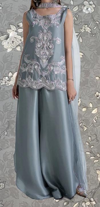 A woman wearing a gray dress with intricate embroidery and a matching gray skirt, standing against a floral patterned background.