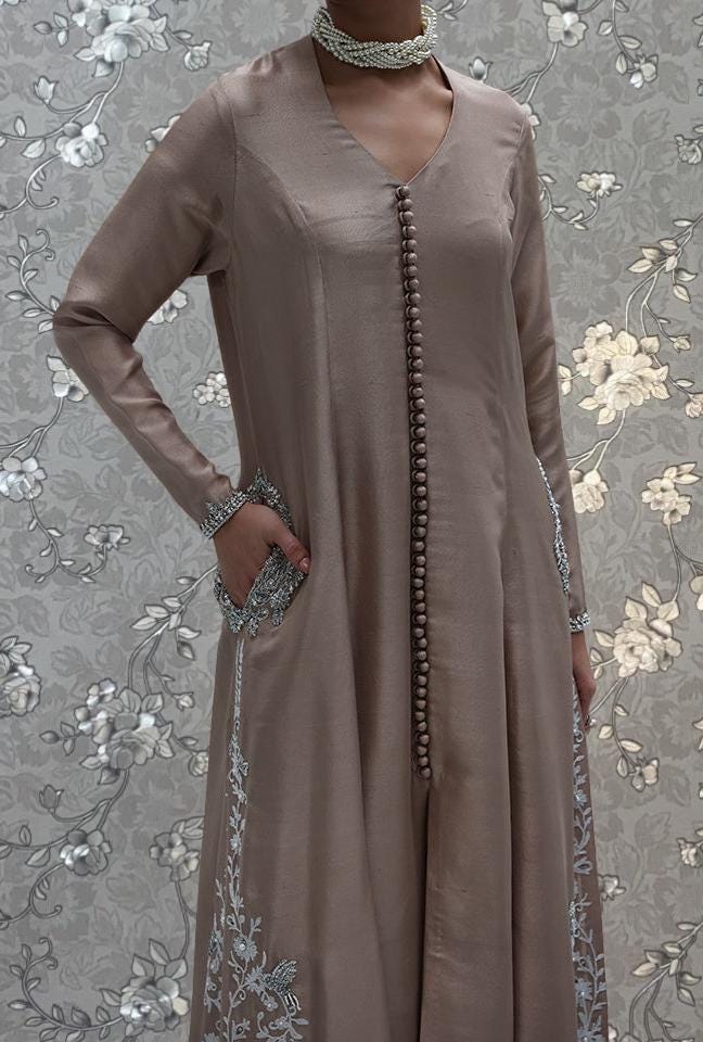 A woman wearing a long, beige dress with intricate embroidery and a choker necklace stands against a floral patterned background.