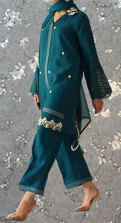 A person wearing a teal green dress with lace sleeves and gold accents, standing against a gray floral background.