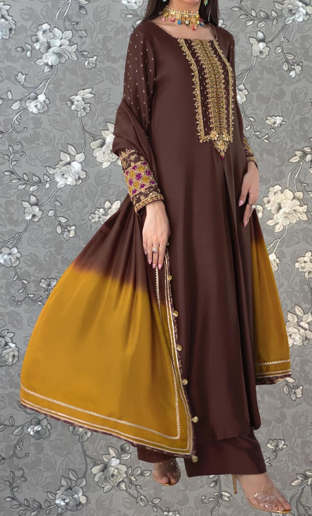 A woman wearing a brown and gold dress with a gold and yellow patterned shawl, standing against a gray floral background.