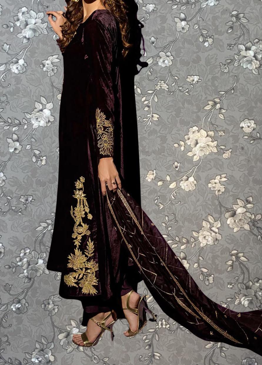 A woman wearing a long, burgundy dress with gold embroidery stands against a gray floral background.