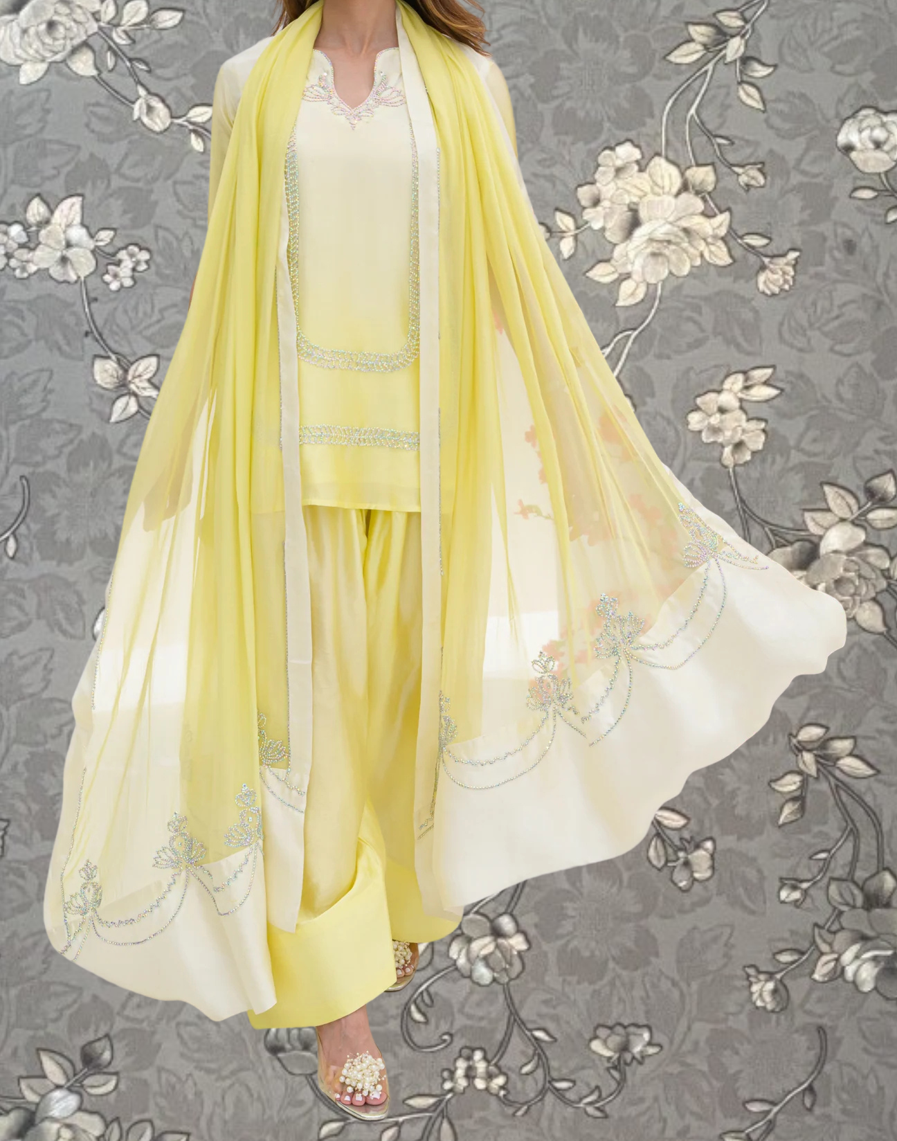 A woman wearing a yellow dress with a long, flowing yellow shawl, standing against a gray floral background.